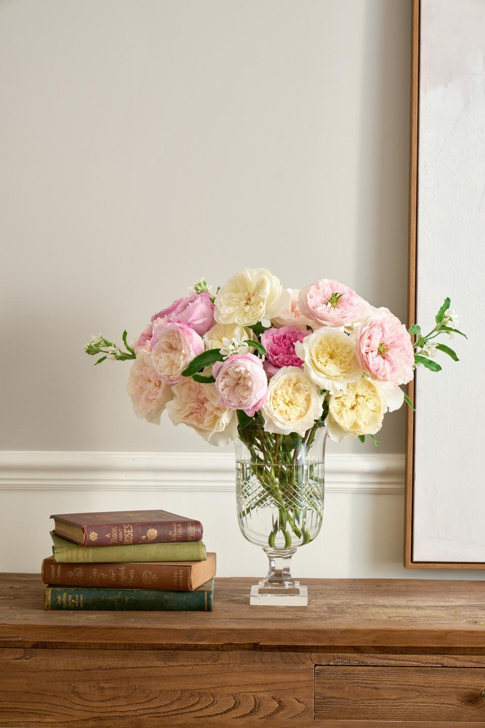 A bouquet of pink, cream, and white roses in a clear glass vase, styled on a wooden console table next to a stack of vintage books against a neutral wall.