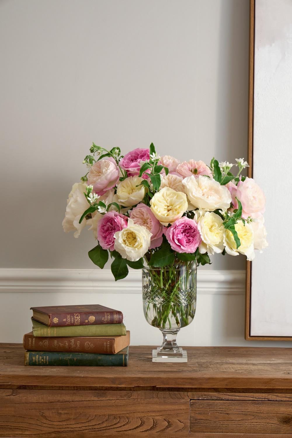 A bouquet of pink, cream, and white roses arranged in a clear glass vase on a wooden table, with a stack of vintage books beside it against a neutral wall.