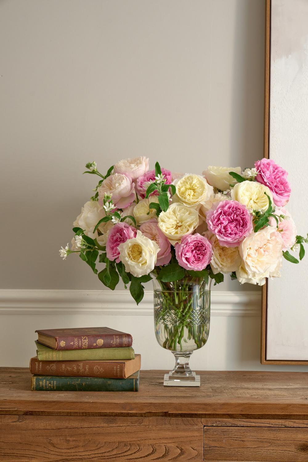 A bouquet of pink, cream, and white roses in a clear glass vase, styled on a wooden console table next to a stack of vintage books against a neutral wall.