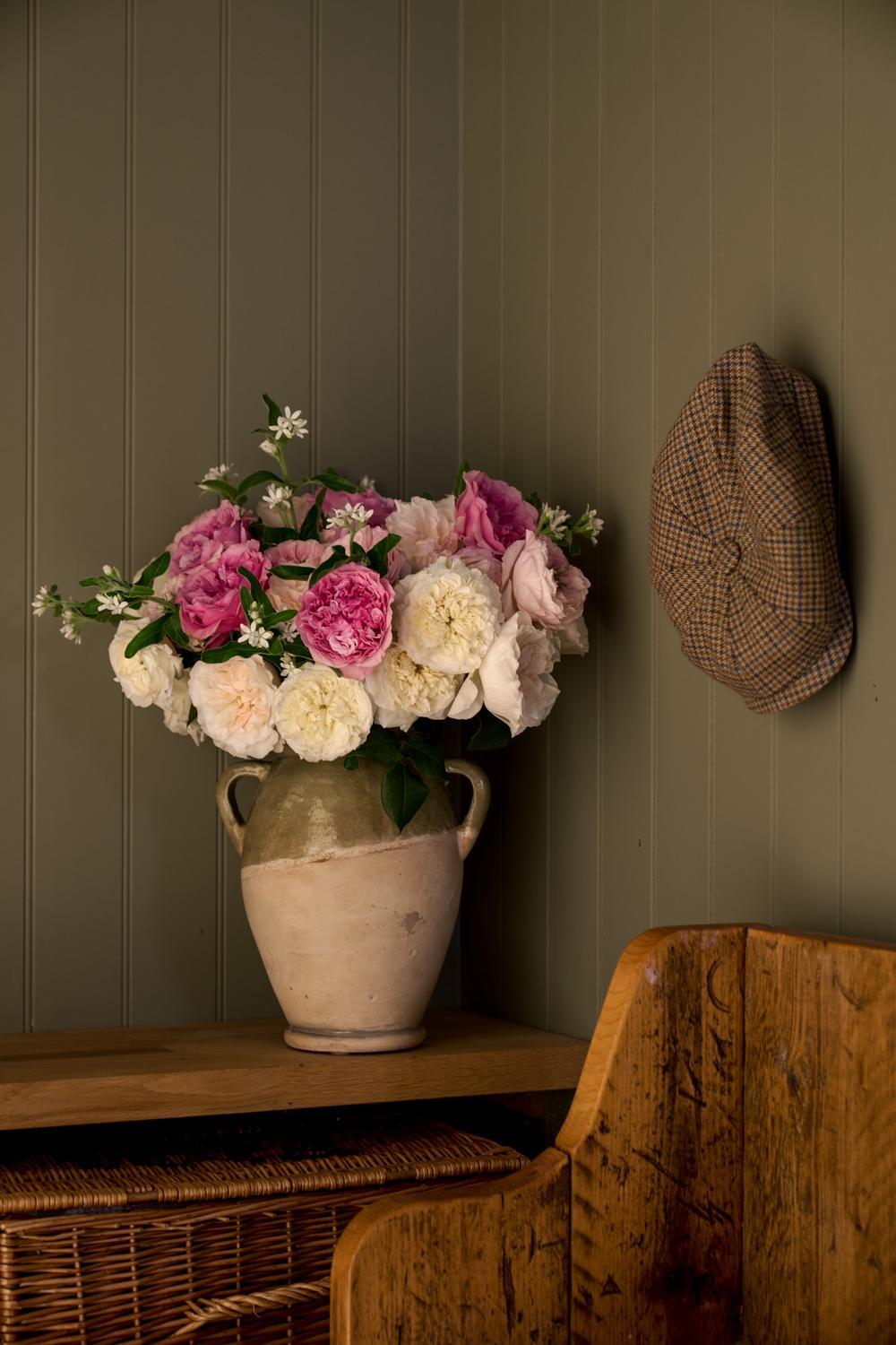 A rustic ceramic vase filled with pink and white roses and small white flowers, placed on a wooden table against a dark olive paneled wall. A brown tweed flat cap hangs on the wall to the right, and part of a wooden chair is visible in the foreground.